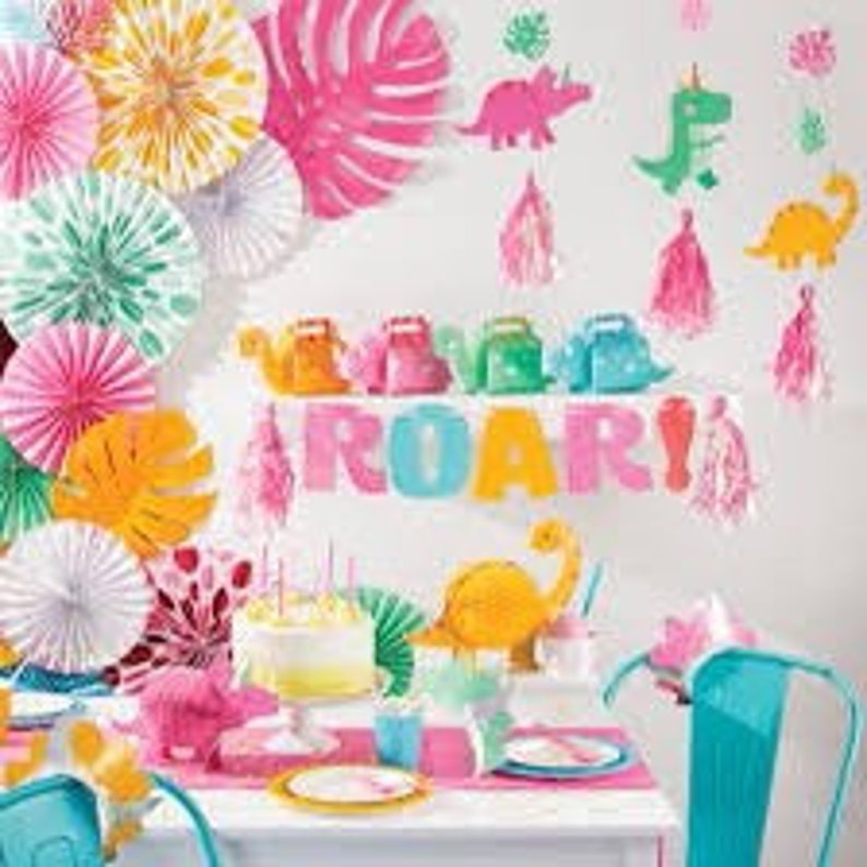 Op de afbeelding: Een dinosaurus-thema feesttafel met groene, roze en gele decoraties. De tafel is gedekt met borden, bekers en een taart. Er zijn ook papieren waaier, dinosaurus-uitknipsels en een banner met de tekst "ROAR!"