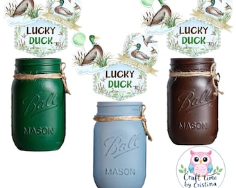 Puede incluir: Tres tarros de cristal, uno verde, uno marrón y uno azul, con una etiqueta impresa que dice "Ball Mason". Cada tarro tiene un lazo de cuerda alrededor de la parte superior. Los tarros están decorados con tapas imprimibles que representan un pato, un globo verde y las palabras "Lucky Duck".