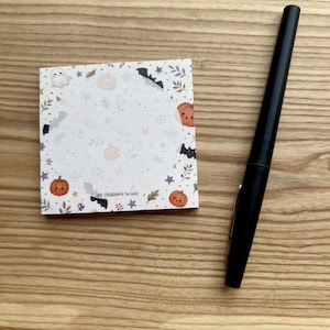 May include: A square notepad featuring a white background with a repeating pattern of small orange pumpkins, grey ghosts, black bats, stars, and foliage, accompanied by a sleek black ballpoint pen with a silver clip on the side.