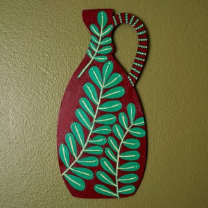 May include: A flat, decorative wooden wall hanging shaped like a pitcher, painted in a deep maroon color. The surface features hand-painted green leaf sprigs with light yellow veins and a striped handle pattern in green and yellow.