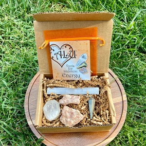 May include: A small cardboard box filled with brown packing paper and crystals. The box contains a clear quartz crystal, a selenite wand, a rose quartz crystal, and a pendulum. The box is sitting on a wooden surface. The box has a small orange bag with the text "Alsu Tools for Intention Energy" on it.