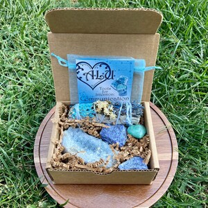 May include: A cardboard box with a blue and white bag inside. The bag has the words "Alou Tools for Intention: Communication" printed on it. The box is filled with blue and green crystals and a bundle of sage.