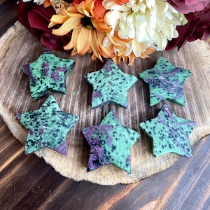 Könnte beinhalten: Sechs sternförmige Steine mit grüner Basis und gesprenkelten lila und schwarzen Mustern. Die Steine sind auf einer Holzoberfläche angeordnet, mit einem Blumenstrauß im Hintergrund. Die Blumen sind eine Mischung aus Orange, Weiß und Burgunderrot.