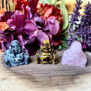 May include: Three small stone figurines of the laughing Buddha, each carved from a different type of stone: grey, tiger's eye, and rose quartz.