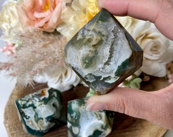 May include: Three green, white, and brown agate crystal points, also known as moss agate, on a wooden surface. The crystals are polished and have a natural, organic look.