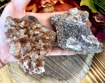 Può includere: Due gruppi di cristalli di calcite marroni, uno con una forma più arrotondata e l'altro con una forma più appuntita, sono esposti su una superficie di legno.
