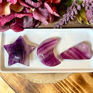 May include: Three purple and white fluorite crystal shapes: a star, a crescent moon, and a cloud. The crystals are arranged on a white rectangular dish.