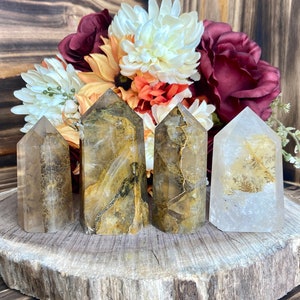 May include: Four clear quartz crystal points with golden inclusions. The points are arranged in a row on a wooden slice. The points are approximately 2 inches tall.
