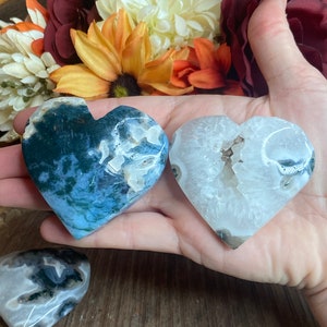 May include: Two heart-shaped agate crystal carvings. One is a light blue and green agate with a white center. The other is a white agate with a clear center and a few dark green spots.