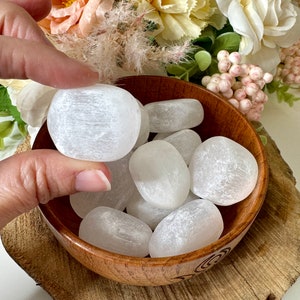 May include: A wooden bowl filled with white selenite tumbled stones. The stones are smooth and have a frosted appearance.