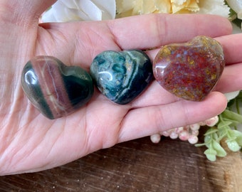 May include: Three heart-shaped stones in green, red, and brown hues are shown. The stones are held in a hand, with a wooden surface and floral arrangement in the background. The stones have unique patterns and textures.