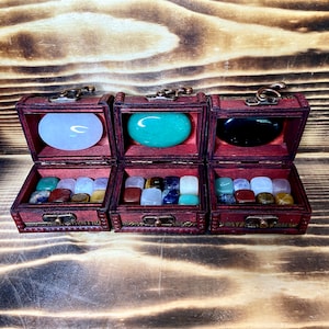 May include: Three wooden treasure chests with hinged lids, each containing a variety of polished gemstones. The chests are lined with velvet and have brass accents. The first chest contains white stones, the second contains green, brown, and blue stones, and the third contains red, brown, and green stones.