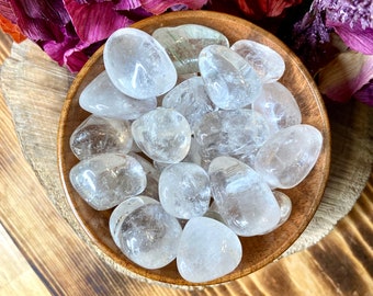 May include: A wooden bowl filled with clear, polished quartz stones. The stones are smooth and rounded, with a translucent appearance. The bowl sits on a wooden surface, with colorful flowers in the background. The stones are a natural, earthy product.