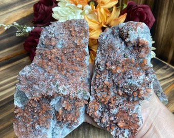 Può includere: Due grappoli di cristalli di quarzo grezzi con inclusioni minerali rosso-arancio. I cristalli sono esposti su una superficie di legno, con uno sfondo di fiori artificiali colorati, tra cui arancione, giallo e bordeaux.