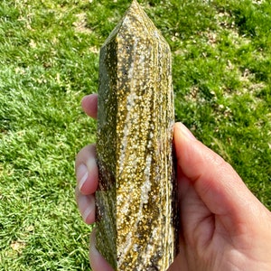 May include: A green and gold crystal point with a unique speckled pattern. The point is smooth and polished, with a pointed tip and a flat base.