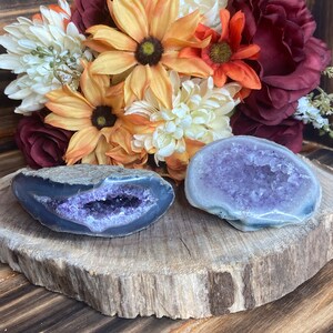 May include: Two amethyst geodes with purple crystals inside. The geodes are on a wooden slice with a bouquet of autumn-coloured flowers behind them.