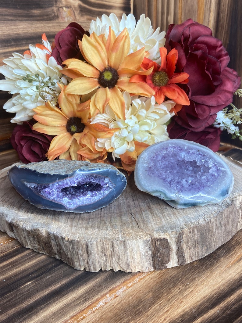 May include: Two amethyst geodes with purple crystals inside. The geodes are on a wooden slice with a bouquet of autumn-coloured flowers behind them.