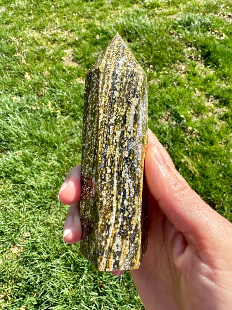 May include: A green and brown crystal point with white flecks. The crystal is pointed at the top and has a flat base.