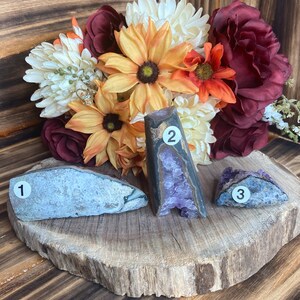May include: Three amethyst geodes, one with a purple interior, one with a grey interior, and one with a blue interior, on a wooden slice. The geodes are numbered 1, 2, and 3.