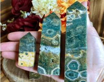 May include: Three green and yellow gemstone obelisks of varying sizes are displayed. The obelisks have a rectangular shape with a pointed top. The gemstones have a unique pattern of green, yellow, and white colors. The obelisks are held in a hand.