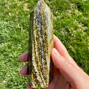 May include: A green and gold crystal point with a unique, swirling pattern. The crystal is polished and has a smooth, glassy finish.