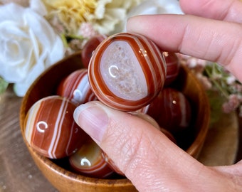 May include: A bowl of carnelian tumbled stones. The stones are a deep red color with white stripes. The stones are smooth and polished.