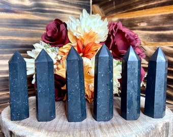 Può includere: Sei torri di cristallo di tormalina nera di diverse altezze sono disposte su una fetta di legno. I cristalli hanno un design sfaccettato e sono in posizione verticale. Sullo sfondo un bouquet di fiori nei toni del rosso, arancione e bianco.