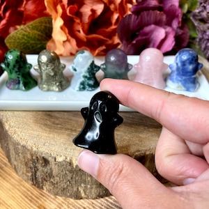 May include: A collection of small, carved ghost figurines made from various types of polished stones. The ghosts are arranged in a row on a white surface. The ghosts are in different colors, including green, gold, white, purple, pink, and blue.