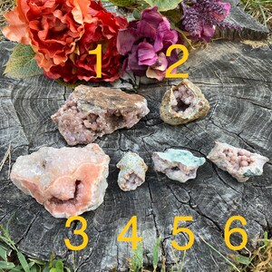 May include: Six pink and white crystal clusters with a variety of shapes and sizes. The crystals are arranged on a wooden surface with numbers 1 through 6 written on the surface.