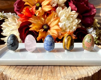 May include: Five polished crystal eggs in various colors, including blue, pink, green, black, and brown, are displayed on a white rectangular tray. The tray is sitting on a wooden surface.