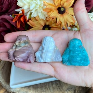 May include: Three small carved stone figurines of a laughing Buddha, each made of a different type of stone: brown, white, and blue.