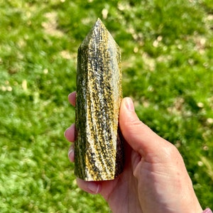 May include: A green and gold striped crystal point, also known as a wand, is held in a hand. The crystal is polished and has a pointed tip.