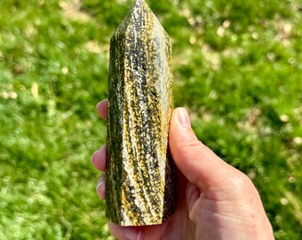 Puede incluir: Una punta de cristal rayada verde y dorada, también conocida como varita, se sostiene en una mano. El cristal está pulido y tiene una punta puntiaguda.