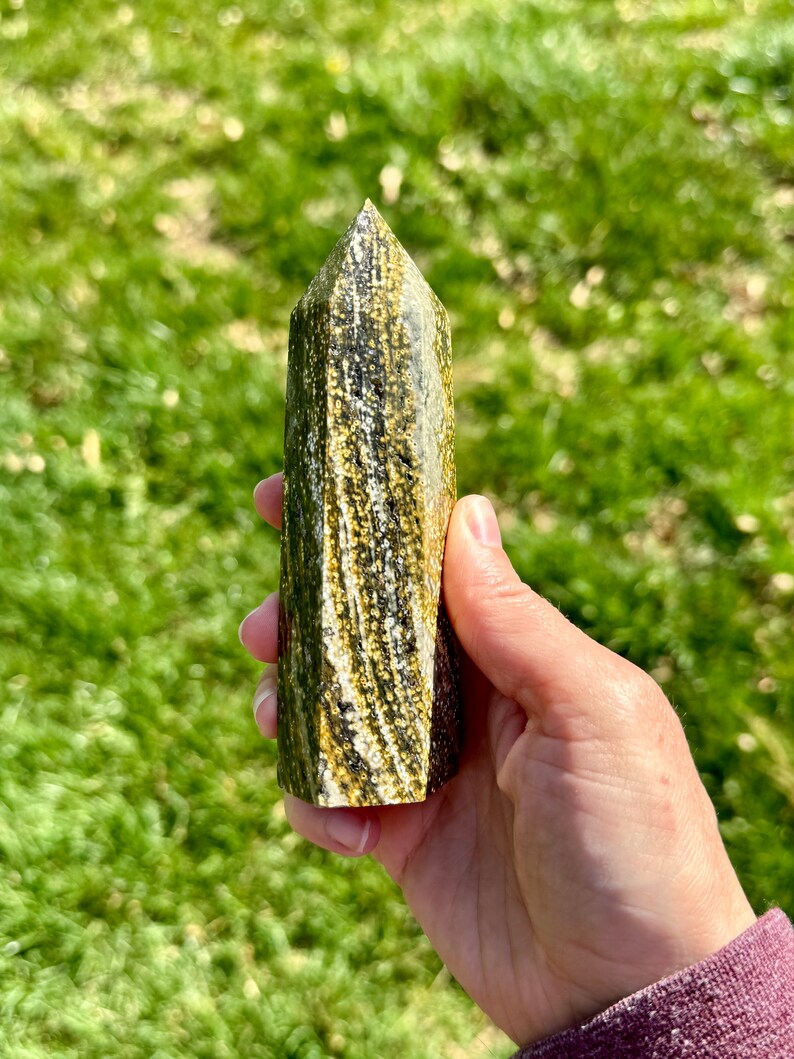 May include: A green and gold striped crystal point, also known as a wand, is held in a hand. The crystal is polished and has a pointed tip.