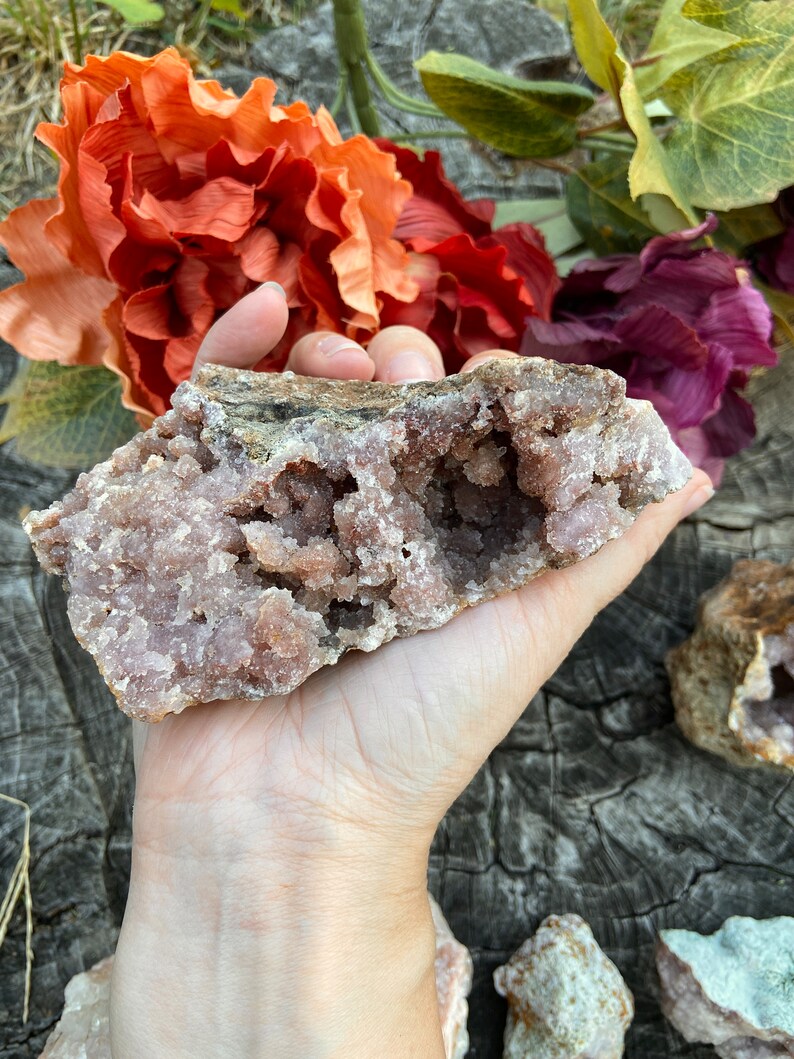 May include: A large, pink amethyst geode with a hollow center and a rough, natural exterior. The geode is being held in a hand, and there are other smaller geodes in the background.