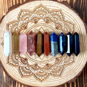 Könnte beinhalten: Ein Set aus acht polierten Kristallstäben in verschiedenen Farben, darunter Weiß, Braun, Rosa, Rot, Orange, Blau und Schwarz. Die Stäbe sind in einem Kreis auf einem Holzsockel mit einem geschnitzten Mandala-Design angeordnet.