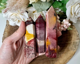 May include: Three polished red, yellow, and white striped crystal points. The points are arranged in a row and are held in a hand. The points are sitting on a wooden surface.