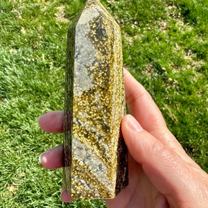 May include: A yellow, brown, and white crystal point with a speckled pattern. The crystal is about 5 inches tall and is held in a hand.