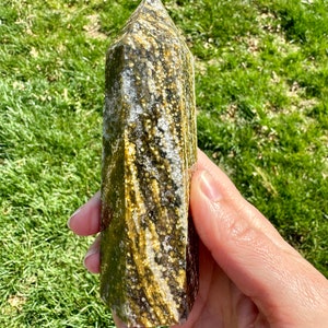 May include: A green and gold crystal point with a unique swirling pattern. The crystal is polished and has a smooth, glassy finish.
