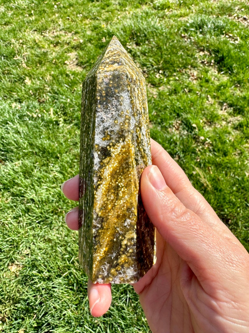 May include: A green and gold crystal point with a unique speckled pattern. The crystal is polished and has a smooth, glassy finish.