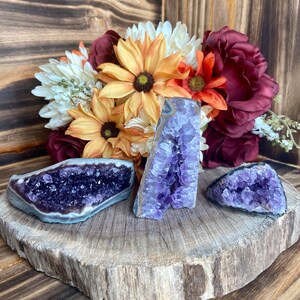 May include: Three amethyst crystal clusters on a wooden slice. The crystals are purple and have a natural, rough texture. The wooden slice is brown and has a natural, rustic look.