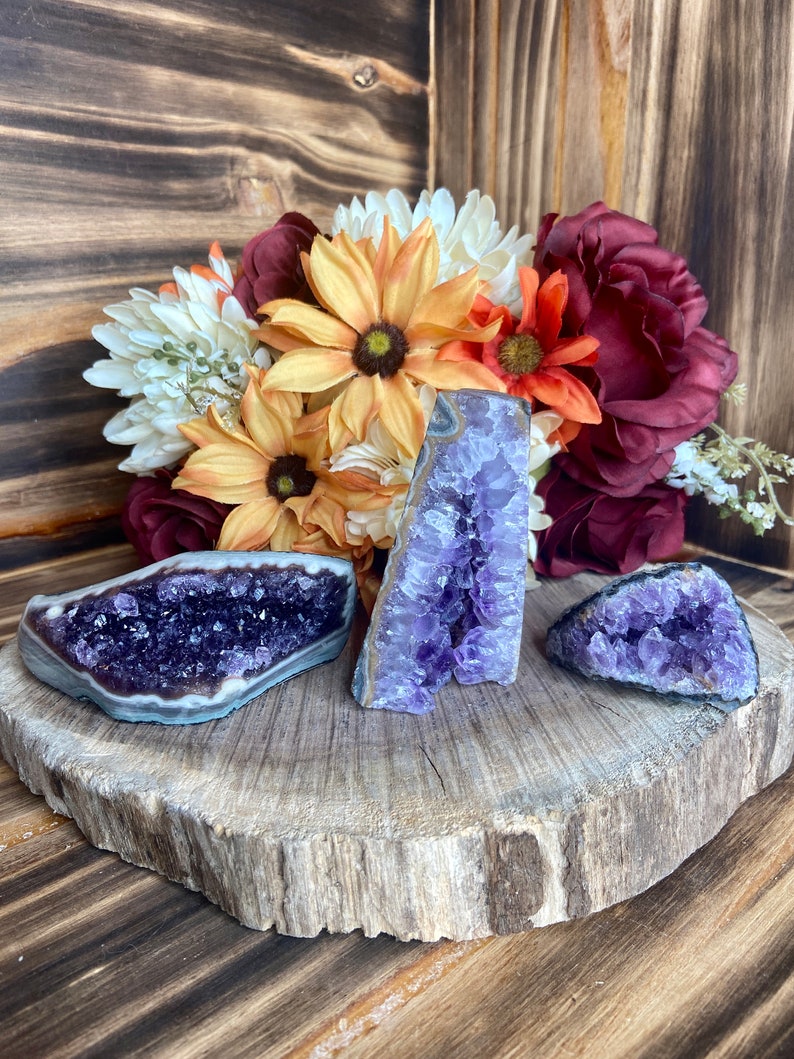 May include: Three amethyst crystal clusters on a wooden slice. The crystals are purple and have a natural, rough texture. The wooden slice is brown and has a natural, rustic look.