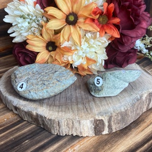 May include: Two smooth, grey stones with the numbers "4" and "5" written on them. The stones are sitting on a wooden slice with a bouquet of autumn-coloured flowers behind them.