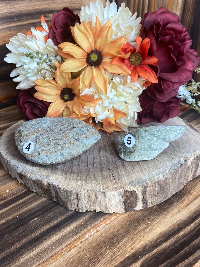 May include: Two smooth, grey stones with the numbers "4" and "5" written on them. The stones are sitting on a wooden slice with a bouquet of autumn-coloured flowers behind them.