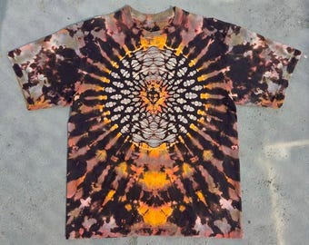 May include: A short-sleeve t-shirt featuring a tie-dye pattern in black, orange, and muted brown tones. The design centers on a symmetrical, intricate mandala-like graphic with radiating lines that extend outward toward the edges of the garment.