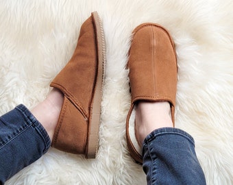 May include: A pair of brown suede slippers with a soft, fuzzy lining. The slippers are worn by a person wearing blue jeans and are sitting on a white fluffy rug.