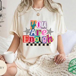 May include: Ivory-colored t-shirt with the text "You Are Enough" in colorful, block-letter style. The design includes star and checkered accents. The shirt is a casual, oversized fit.