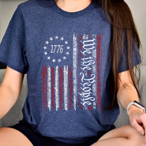 May include: A navy blue t-shirt with a distressed American flag design. The flag has red and white stripes and a circle of stars with the year 1776 in the center. The text "We the People" is written vertically on the right side of the flag.