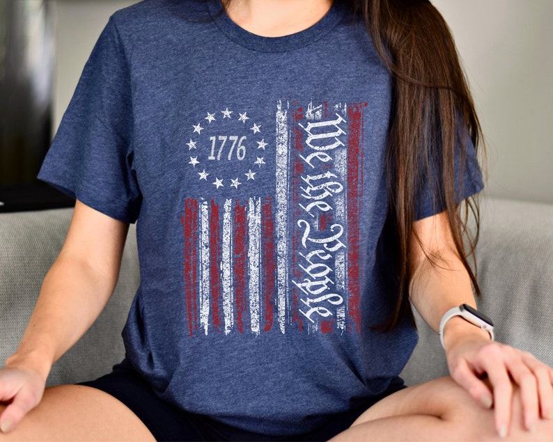 May include: A navy blue t-shirt with a distressed American flag design. The flag has red and white stripes and a circle of stars with the year 1776 in the center. The text "We the People" is written vertically on the right side of the flag.