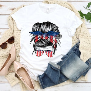 May include: White t-shirt featuring a graphic of a woman with a messy bun and a red, white and blue bandana. The bandana has a splatter paint design.